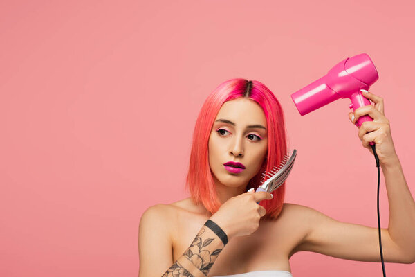 tattooed young woman brushing colorful hair while holding hair dryer isolated on pink 