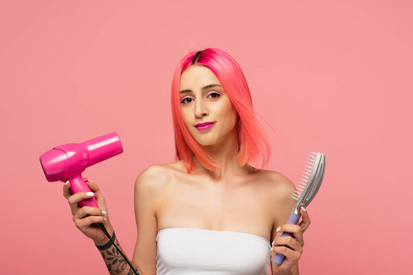 positive young woman with colorful hair holding hair brush and dryer isolated on pink 