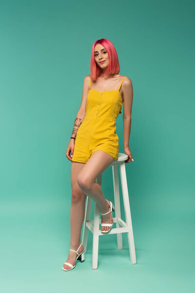 full length of happy young woman with pink dyed hair sitting on stool on blue 