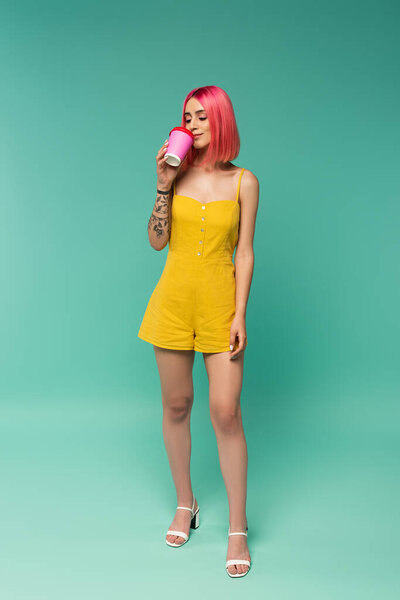 full length of young woman with pink dyed hair holding paper cup while drinking coffee to go on blue 