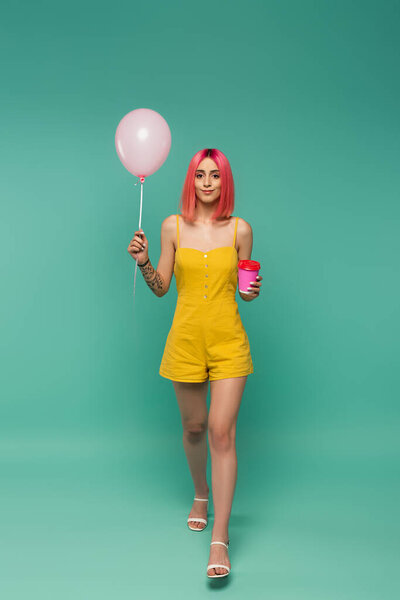 full length of young woman with pink dyed hair holding paper cup and balloon on blue 
