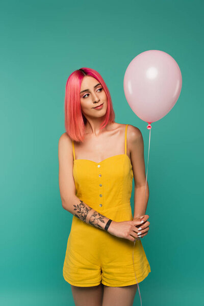 curious young woman with pink dyed hair looking at balloon isolated on blue 