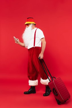 Noel Baba güneş gözlüklü, akıllı telefon kullanıyor ve kırmızı arka planda bavulu tutuyor.