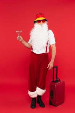 Güneş gözlüklü Noel Baba kırmızı arka planda bavulun yanında kokteyl tutuyor.