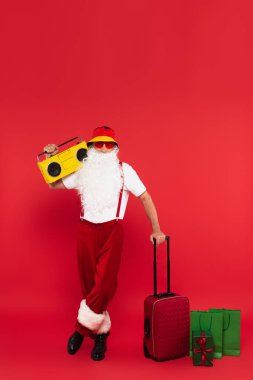 Güneş gözlüklü Noel Baba bavulun yanında teyp taşıyor ve kırmızı arka planda alışveriş torbaları var.