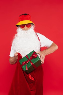 Panama ve güneş gözlüklü Noel Baba kırmızı üzerine izole edilmiş yay ve çuvalla hediye koyar. 