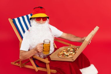 Noel Baba güneş gözlüklü, elinde pizza kutusu ve kırmızı arka planda bir bardak bira tutuyor.