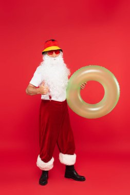 Güneş gözlüklü Noel Baba elinde yüzme yüzüğü ve kırmızı arka planda beliriyor.