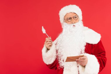 Kırmızı üzerine izole edilmiş kalem ve zarf tutan düşünceli Noel Baba. 