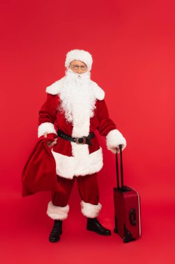 Noel Baba kırmızı arka planda çanta ve çuval tutuyor.