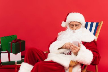 Noel Baba güvertedeki sandalyede akıllı telefon kullanıyor. Kırmızı arka planda kızak üstünde hediyeler.