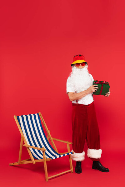 Santa claus in sunglasses holding present near deck chair on red background