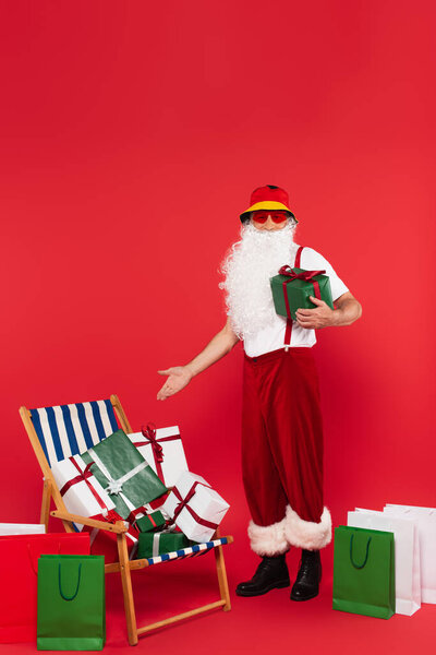 Santa claus in sunglasses pointing at presents on deck chair near shopping bags on red background