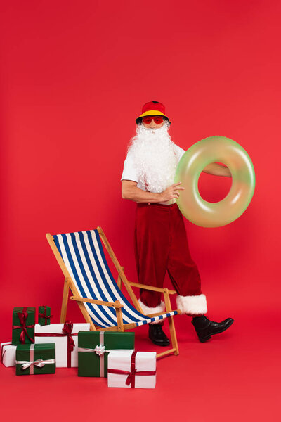Santa claus in sunglasses holding swim ring near deck chair and presents on red background