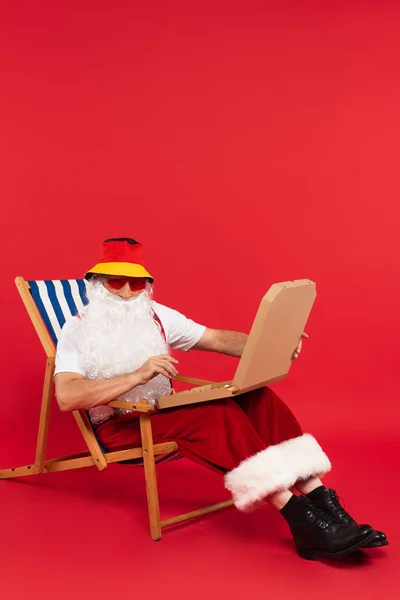 穿着睡衣的圣诞老人 戴着太阳镜 把披萨盒放在红色背景的餐椅上 — 图库照片