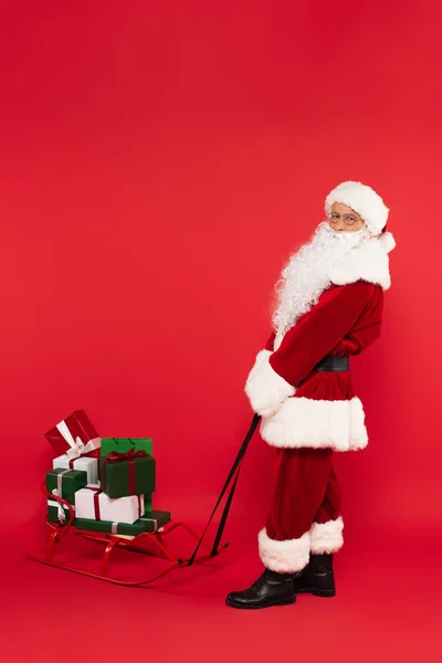 Weihnachtsmann Zieht Schlitten Mit Geschenken Auf Rotem Hintergrund — Stockfoto