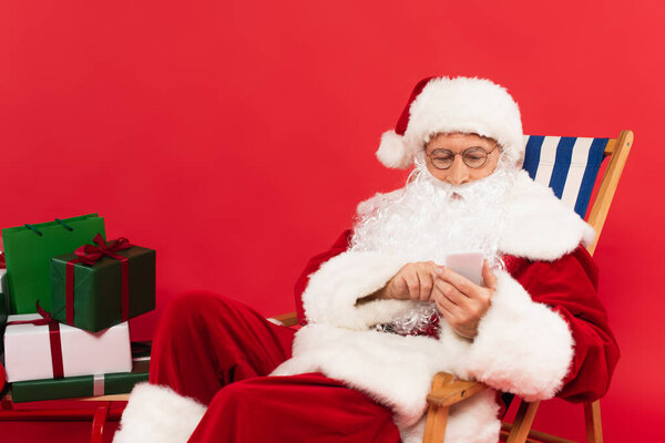 Santa using smartphone on deck chair near presents on sleigh on red background