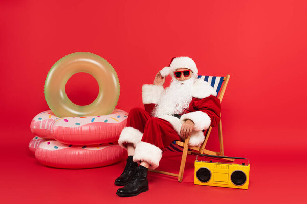 Santa claus in sunglasses sitting on deck chair near boombox and swim rings on red background