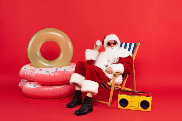 Santa claus in sunglasses and costume sitting on deck chair near swim rings and boombox on red background