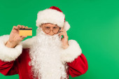 Santa Claus mluví na smartphone a drží kreditní kartu izolované na zelené