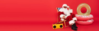 Güneş gözlüklü Noel Baba güvertedeki sandalyede klasik müzik kutusunun yanında oturuyor ve kırmızı arka planda yüzen yüzükler, afiş 