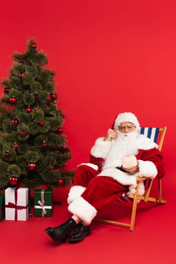 Noel Baba, kırmızı arka planda Noel ağacının yanında hindistan cevizli kokteyl tutuyor.