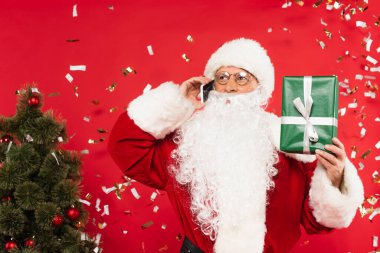 Noel Baba akıllı telefondan konuşuyor ve kırmızı arka planda Noel ağacının yanında konfeti altında hediyeyi tutuyor.