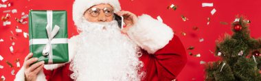 Noel Baba akıllı telefondan konuşuyor ve hediyesini konfeti yakınında tutuyor ve kırmızı arka planda Noel ağacı, afiş