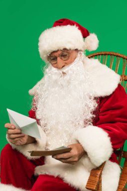 Gözlüklü Noel Baba, hasır sandalyede yeşilde izole edilmiş zarflar tutuyor.