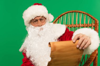Gözlüklü Noel Baba, yeşil renkli hasır sandalyede bulanık kağıt tutuyor.