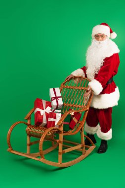 Noel Baba yeşil arka planda sallanan sandalyede hediyeleri gösteriyor.
