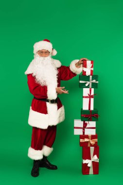 Gözlüklü ve kostümlü Noel Baba yeşil arka plandaki hediye kutularını gösteriyor. 