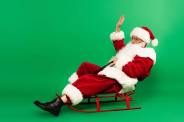 Noel Baba yeşil arka planda kızakta otururken el sallıyor.