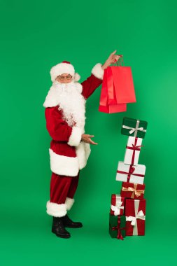 Noel Baba kostümlü hediyeleri gösteriyor ve yeşil arka planda alışveriş torbaları tutuyor.