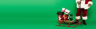 Cropped view of santa claus in costume standing near presents on sleigh on green background, banner  clipart