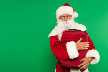 Şapkalı ve gözlüklü Noel Baba yeşilde izole edilmiş bavulu kucaklıyor.