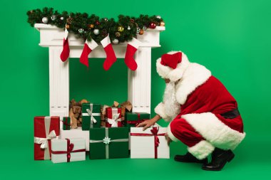 Noel Baba, yeşil arka planda Noel çoraplarıyla süslü şöminenin yanına hediye koyuyor.