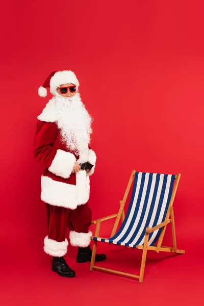 Papai Noel Traje Óculos Sol Perto Cadeira Deck Fundo Vermelho — Fotografia de Stock