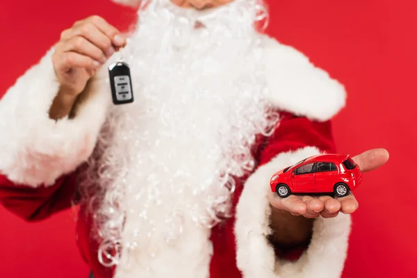 Cropped view of car model on hand of blurred santa with key isolated on red