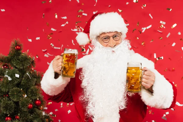 圣诞老人在红底的五彩纸屑和圣诞树旁举着一杯啤酒 — 图库照片