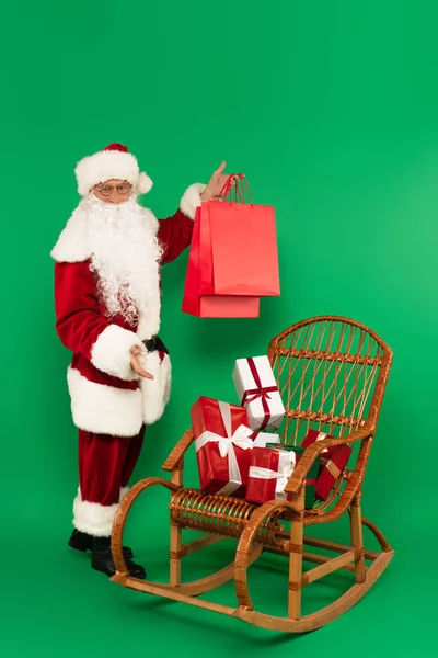 Weihnachtsmann Kostüm Hält Einkaufstüten Neben Geschenken Auf Korbsessel Auf Grünem — Stockfoto