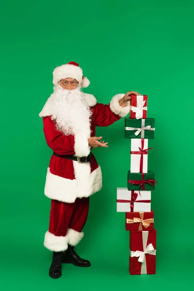 Święty Mikołaj Okularach Kostiumie Wskazując Ułożone Pudełka Zielonym Tle — Zdjęcie stockowe