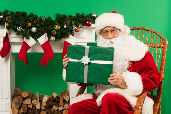 Santa claus holding present on rocking chair near fireplace on green background 