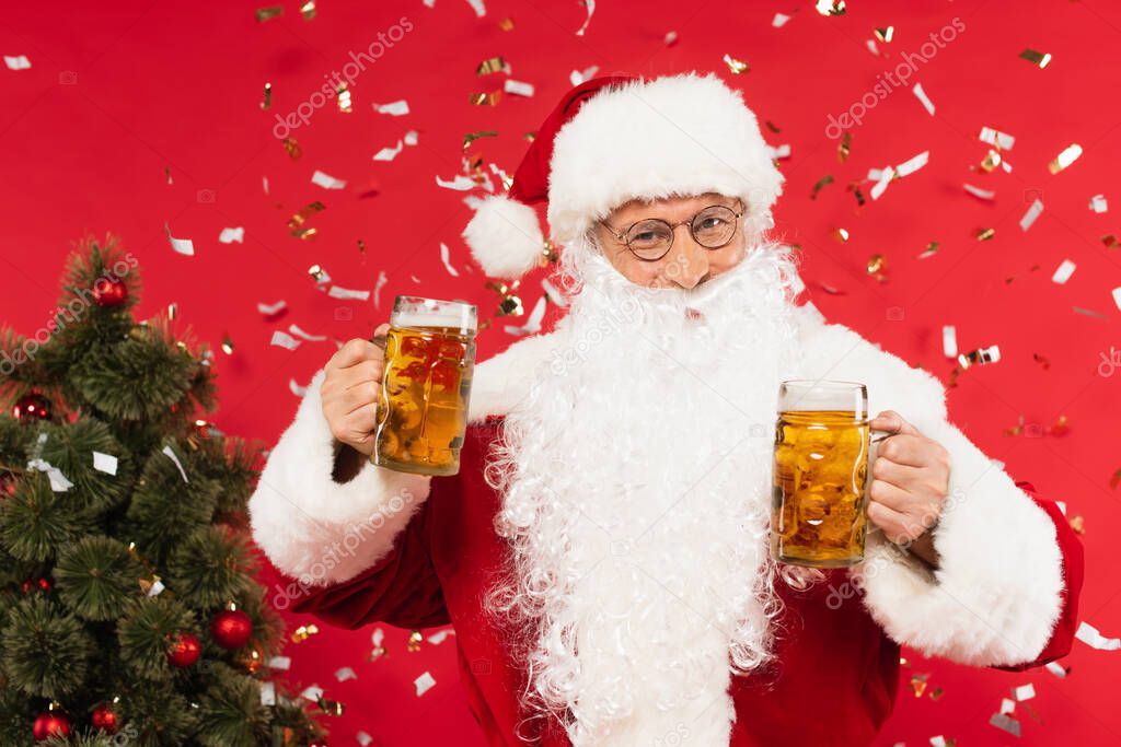 Santa claus holding glasses of beer near confetti and christmas tree on red background