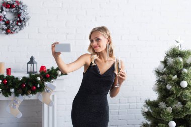 Şampanya bardağıyla mutlu genç bir kadın yılbaşı ağacının yanındaki akıllı telefondan selfie çekiyor.