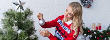 Süslü kırmızı kazaklı sarışın kadın evde Noel ağacı süslüyor, afiş.
