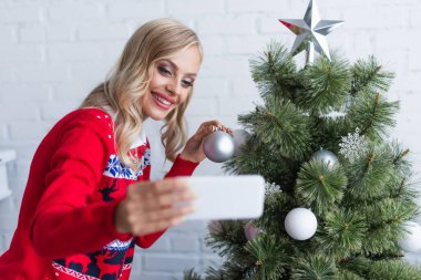 Sıcak kazaklı neşeli sarışın kadın evde Noel ağacının yanında selfie çekiyor, bulanık ön plan.