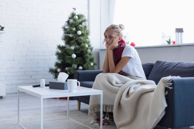 Bulanık Noel ağacının yanındaki sıcak battaniyenin altında kanepede oturan hasta bir kadın.