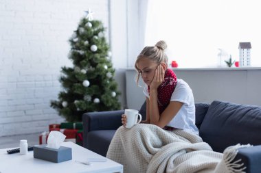 Hasta ruhlu bir kadın, bulanık arka planda bir bardak sıcak içecekle Noel ağacının yanında oturuyor.