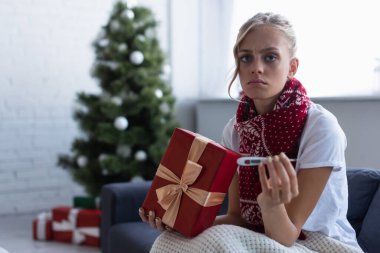 Hediye kutusu ve termometresi olan üzgün kadın Noel ağacının yakınındaki kameraya bakıyor ve bulanık arkaplanda hediyeler sunuyor.